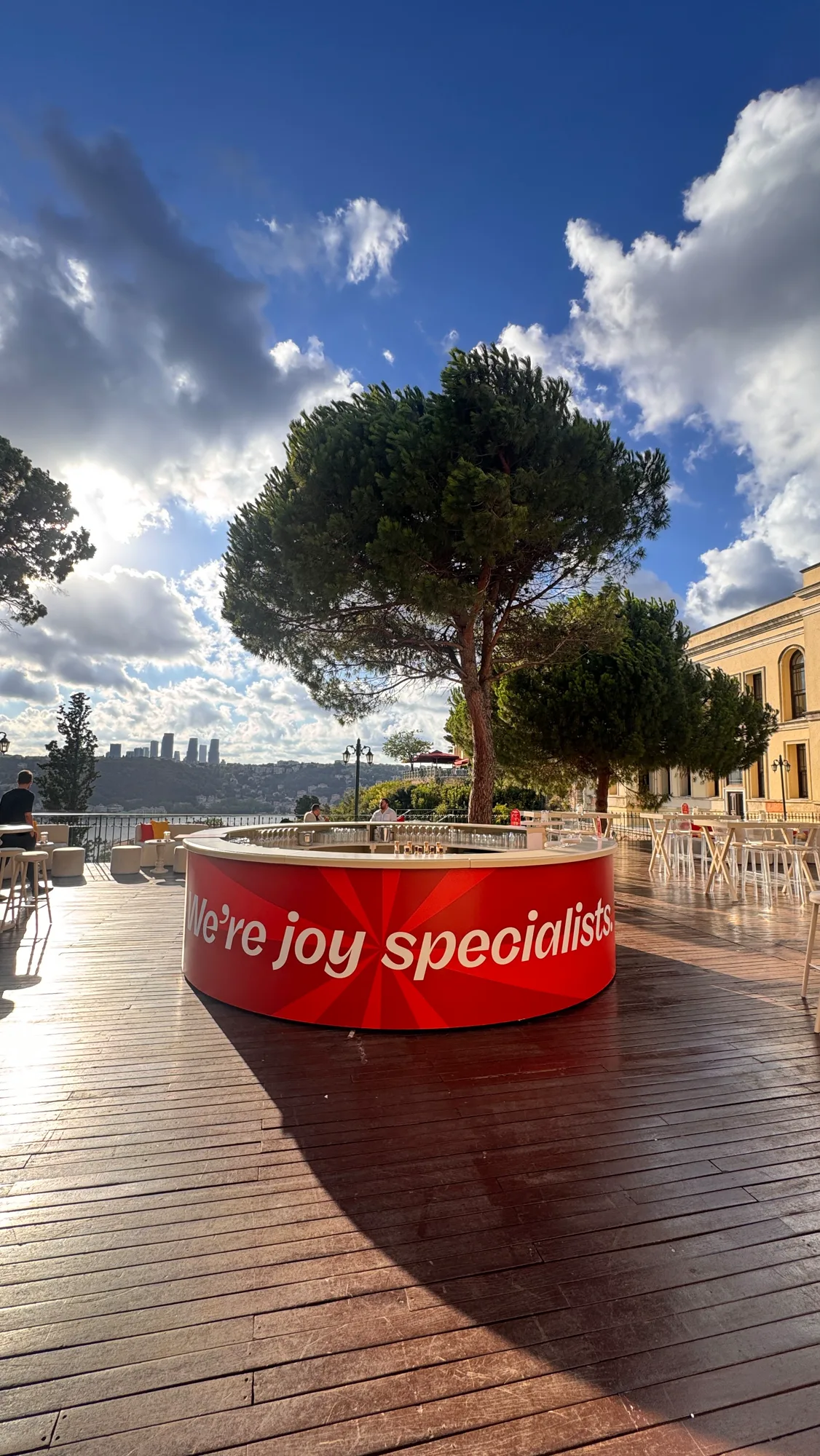 Yuvarlak kırmızı bar: “We’re joy specialists” yazısı, arkada çam ve Boğaz