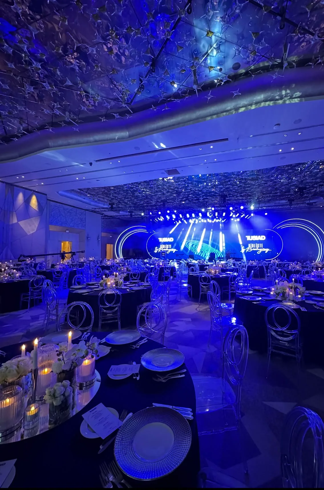 Aynalı tavan altında mavi ışıkla yıkanmış salon, sahnede TÜSİAD ekranı