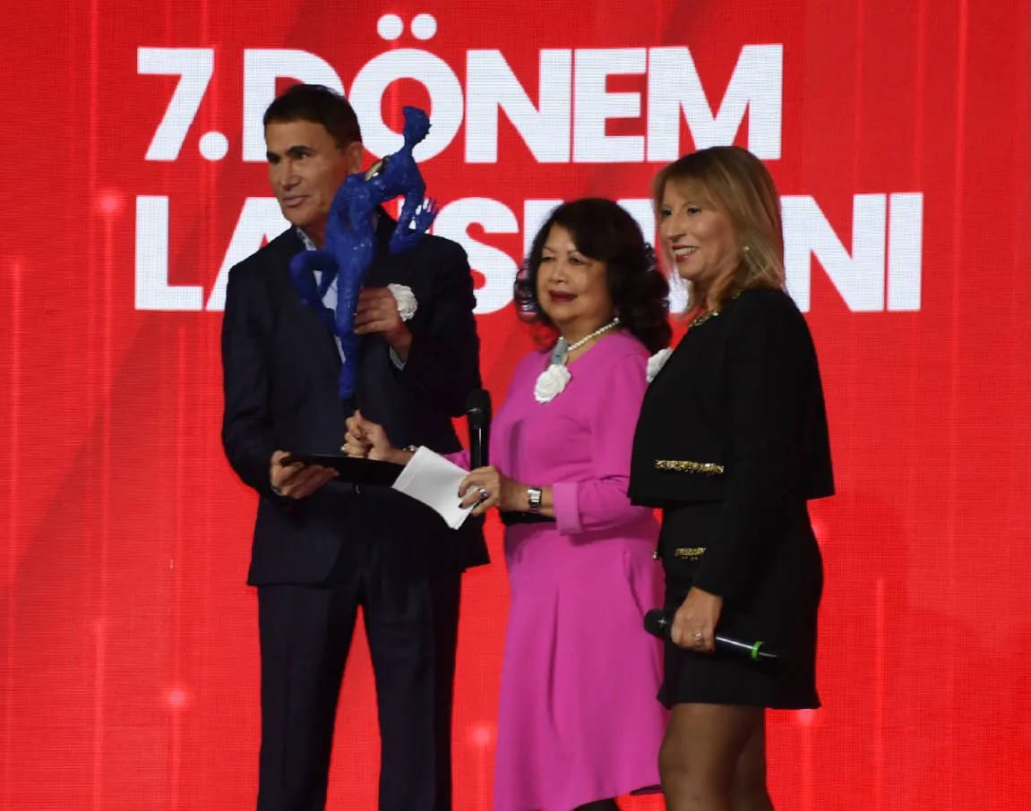Sahnede takdim anı: kırmızı “7. Dönem Lansmanı” ekranı önünde ödül objesi